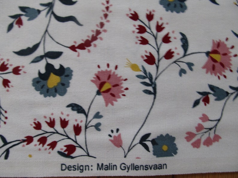 Op de afbeelding: Witte stof met een bloemenpatroon. Het patroon bevat roze, rode, blauwe en gele bloemen en groene bladeren. De tekst "Design: Malin Gyllensvaan" is zichtbaar.