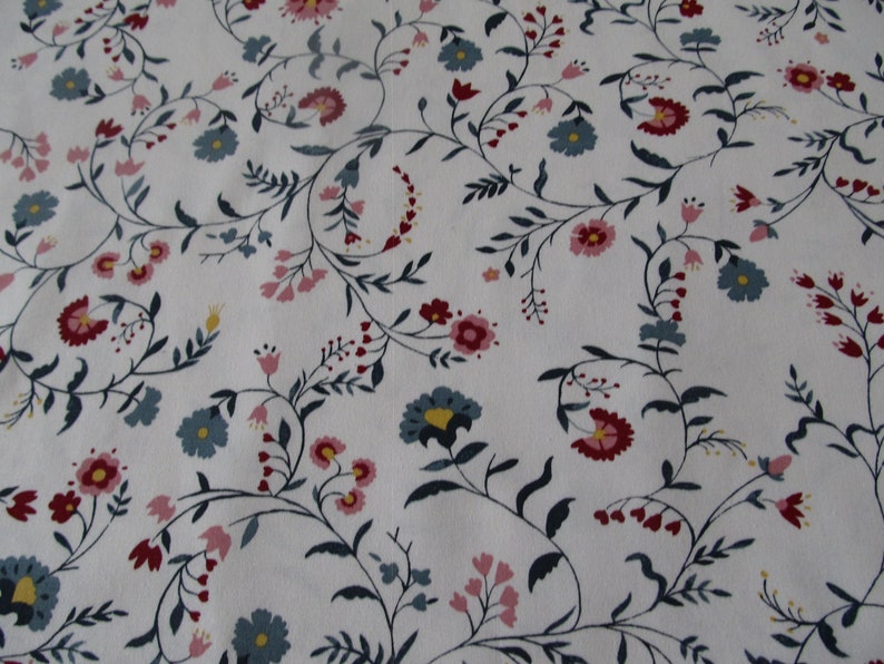 Op de afbeelding: Witte stof met een bloemenpatroon. Het ontwerp toont kronkelende stelen met kleine bladeren en clusters van bloemen in roze, rood en blauw. De algehele esthetiek is delicaat en vintage, doet denken aan een klassieke print.