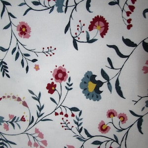 Op de afbeelding: Witte stof met een herhalend bloemenpatroon. Het ontwerp toont gestileerde bloemen in roze, rood en blauw, met gele centra en donkergroene bladeren en stelen. De algehele esthetiek is delicaat en vrouwelijk.