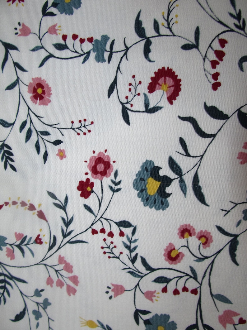 Op de afbeelding: Witte stof met een herhalend bloemenpatroon. Het ontwerp toont gestileerde bloemen in roze, rood en blauw, met gele centra en donkergroene bladeren en stelen. De algehele esthetiek is delicaat en vrouwelijk.