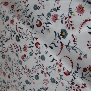 Op de afbeelding: Witte stof met een herhalend bloemenpatroon. Het patroon bevat roze, blauwe en rode bloemen, groene bladeren en donkere stelen. De stof is gevouwen, waardoor de textuur en de manier waarop het drapeert zichtbaar zijn. Het ontwerp cre&euml;ert een vloeiende, organische esthetiek.