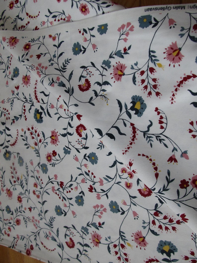 Op de afbeelding: Witte stof met een herhalend bloemenpatroon. Het patroon bevat roze, blauwe en rode bloemen, groene bladeren en donkere stelen. De stof is gevouwen, waardoor de textuur en de manier waarop het drapeert zichtbaar zijn. Het ontwerp cre&euml;ert een vloeiende, organische esthetiek.