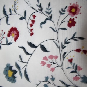 Op de afbeelding: Witte stof met een bloemenpatroon. Het ontwerp toont gestileerde bloemen in tinten rood, roze en blauwgroen, met gele centra en donkergrijze stelen en bladeren. Het geheel geeft een delicaat, botanisch effect.