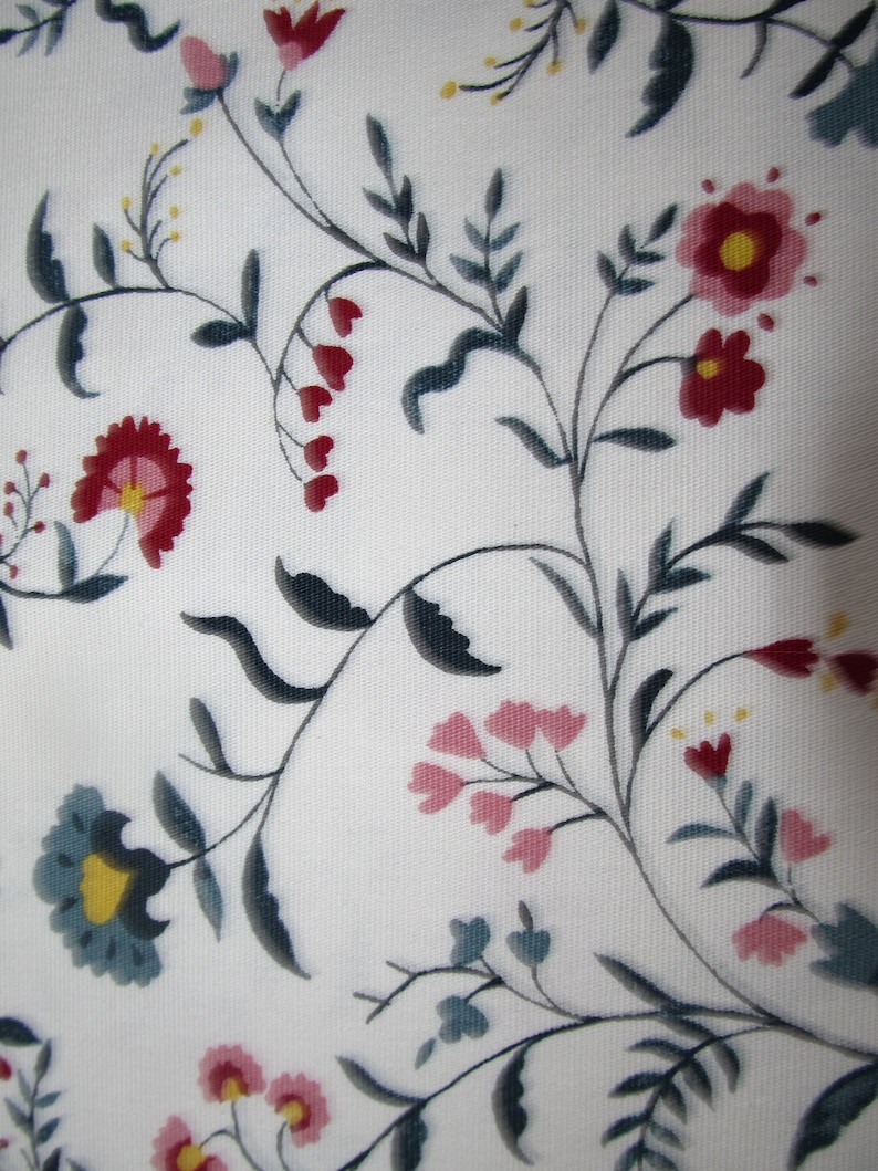 Op de afbeelding: Witte stof met een bloemenpatroon. Het ontwerp toont gestileerde bloemen in tinten rood, roze en blauwgroen, met gele centra en donkergrijze stelen en bladeren. Het geheel geeft een delicaat, botanisch effect.