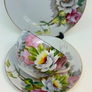 Op de afbeelding: Een vintage theekopje en schotelset met een bloemenontwerp. Het kopje en de schotel zijn wit met een delicaat bloemenpatroon in tinten roze, wit en groen. De bloemen zijn realistisch geschilderd en hebben een licht vervaagde uitstraling. De set heeft een gouden rand rond de randen.