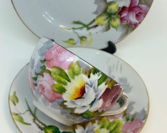 May include: A vintage teacup and saucer set with a floral design. The cup and saucer are white with a delicate floral pattern in shades of pink, white, and green. The flowers are painted in a realistic style and have a slightly faded look. The set has a gold trim around the edges.