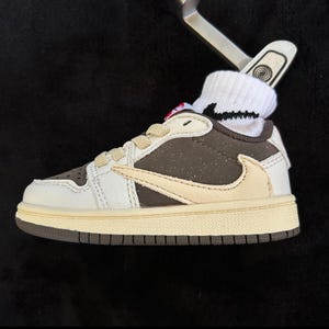 Könnte beinhalten: Ein Miniatur-Sneaker in Braun, Creme und Weiß mit einem cremefarbenen Nike-Swoosh. Der Schuh hat cremefarbene Schnürsenkel und eine braune Sohle. Eine weiße Socke mit schwarzen Akzenten befindet sich im Schuh, und ein silberner Golfschläger ist in die Socke gesteckt.