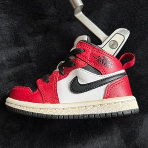 Puede incluir: Una zapatilla Air Jordan 1 roja, blanca y negra para bebés. El zapato presenta una parte superior de cuero rojo, cordones negros, un panel lateral blanco con un logotipo de Nike negro y una suela negra y blanca. El logotipo de Air Jordan es visible en el lateral.
