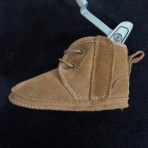 Puede incluir: Una bota de bebé de ante marrón claro con un diseño de cordones y una lengüeta en la parte trasera. La bota está sobre un fondo oscuro y tiene una suela cosida. Un putter de golf plateado está insertado en la bota.