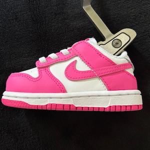May include: A white and pink baby sneaker with a velcro strap. The sneaker features a pink Nike swoosh and a white sole. The shoe is approximately 10 cm long.
