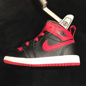 Puede incluir: Una zapatilla Air Jordan 1 en miniatura, negra y roja, con suela blanca y detalles rojos. El icónico logotipo de Nike es visible en el lateral. La zapatilla es un objeto de colección, posiblemente una pieza decorativa o un juguete.