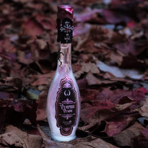 Può includere: Una bottiglia di vetro trasparente con un'etichetta scura che recita "Vampire Venom". La bottiglia ha un tappo argentato con un gioiello rosso e un ciondolo argentato appeso. La bottiglia è appoggiata su un letto di foglie marroni.