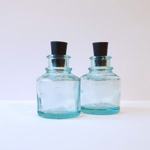 May include: Two clear glass bottles with black stoppers. The bottles are textured and have a light blue tint.