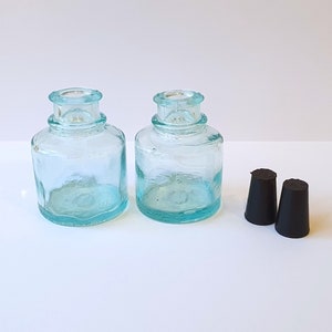 May include: Two clear glass bottles with black rubber stoppers. The bottles are a light blue colour and have a rounded shape.