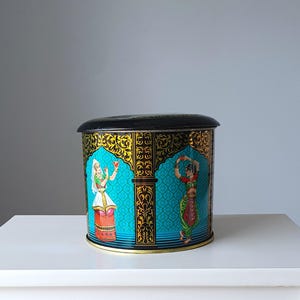 Puede incluir: Un recipiente redondo de hojalata negro y dorado con un fondo azul y un diseño de dos mujeres con ropa tradicional india. Las mujeres están representadas bailando y sosteniendo una taza.