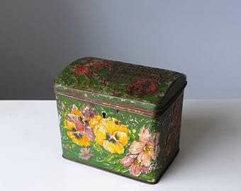 May include: Antique green metal storage tin with a curved lid. The tin is adorned with hand-painted yellow, pink, and purple pansies. The lid features faded text and a circular emblem. The tin has a keyhole, likely from the early 1900s.