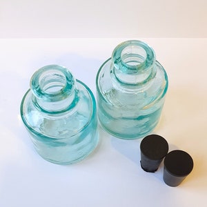 May include: Two clear glass bottles with a light blue tint and black rubber stoppers. The bottles are round and have a wide mouth.