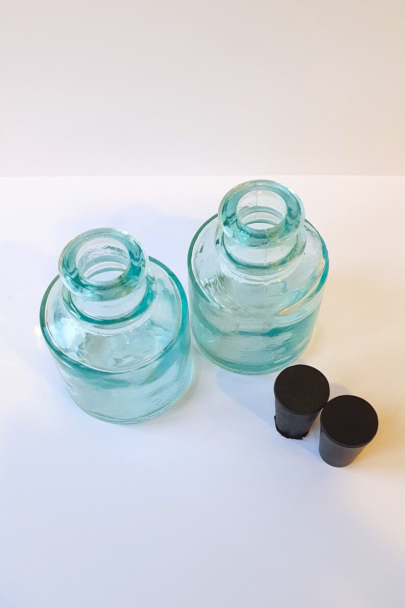May include: Two clear glass bottles with a light blue tint and black rubber stoppers. The bottles are round and have a wide mouth.