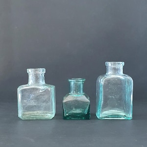 Könnte beinhalten: Drei Glasflaschen, zwei sind klar und eine ist grün. Die Flaschen sind quadratisch und haben einen Vintage-Look.
