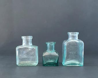 May include: Three glass bottles, two are clear and one is green. The bottles are square shaped and have a vintage look.