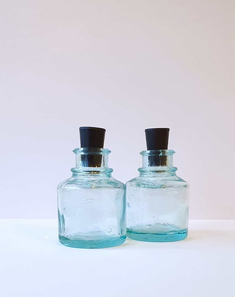 May include: Two clear glass bottles with black stoppers. The bottles are textured and have a light blue tint.