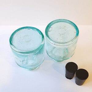 May include: Two clear glass bottles with a light green tint and black rubber stoppers. The bottles are round and have a wide mouth.