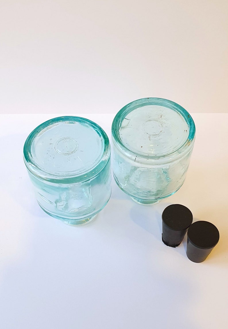 May include: Two clear glass bottles with a light green tint and black rubber stoppers. The bottles are round and have a wide mouth.