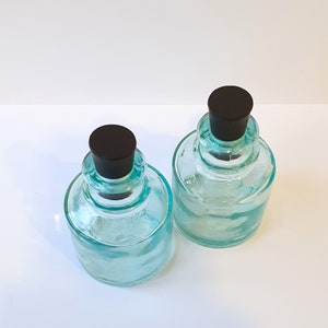 May include: Two clear glass bottles with black lids. The bottles are a light blue colour and have a textured surface.