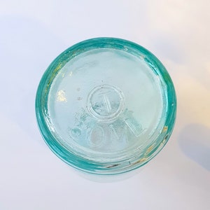 May include: A clear glass bottle with a green rim and a circular embossed mark that reads "COM" with a "T" inside the circle.
