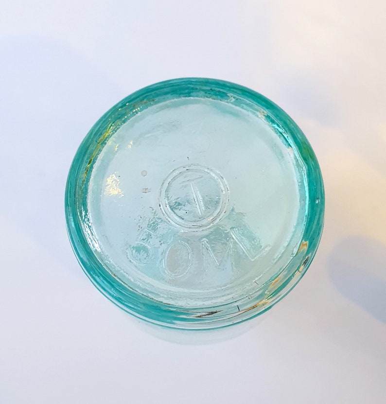 May include: A clear glass bottle with a green rim and a circular embossed mark that reads "COM" with a "T" inside the circle.