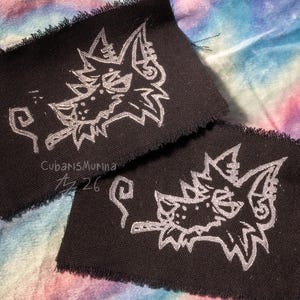 May include: Two rectangular black fabric patches with frayed edges, each featuring a white embroidered graphic of a stylized wolf head smoking a cigarette. The text CubarisMurina and the characters Az 26 are printed in white on the fabric.