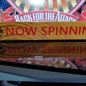 May include: A wooden sign with the words "NOW SPINNING" in red letters. A black and white vinyl record icon is on the left side of the sign.