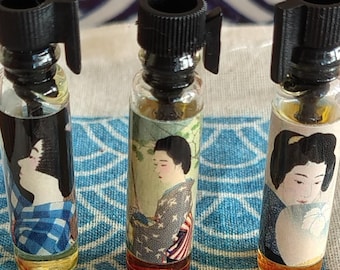 Puede incluir: Tres frascos de vidrio pequeños con tapas de plástico negro, cada uno con un líquido de diferente color. Los frascos tienen etiquetas con ilustraciones de figuras con vestimenta tradicional japonesa sobre una bolsa de tela color crema.