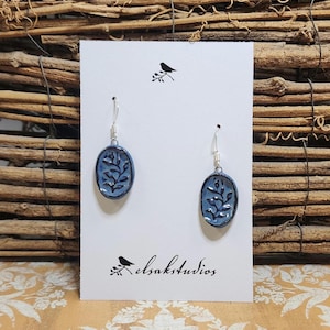 May include: A pair of blue oval earrings with a leaf design, hanging from silver hooks. The earrings are displayed on a white card with a bird illustration and the text "elsakstudios."