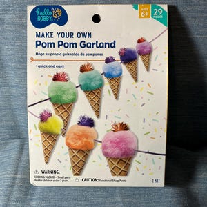 May include: A craft kit for making a pom pom garland. The box features colorful pom poms shaped like ice cream cones. Text on the box reads "MAKE YOUR OWN Pom Pom Garland" and "quick and easy". The kit includes 29 pieces and is for ages 6+.