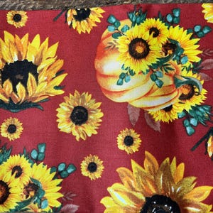 May include: A red fabric featuring a pattern of yellow sunflowers and a pumpkin. The sunflowers have dark brown centers and are surrounded by green leaves. The pumpkin is orange and yellow.