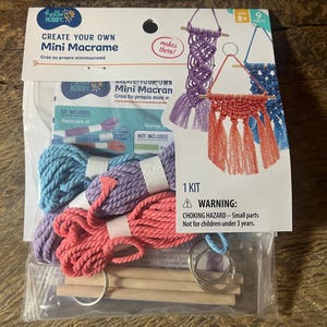 May include: A Hello Hobby Mini Macrame craft kit in a clear plastic package. The kit includes three bundles of cotton cord in blue, purple, and coral, three wooden dowels, and metal keyrings to create three small macrame wall hangings.