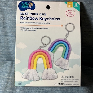 May include: A craft kit for making rainbow keychains. The box features two finished rainbow keychains with colorful embroidery and white fringe. Text on the box reads "MAKE YOUR OWN Rainbow Keychains" and "create up to 2 rainbow keychains".