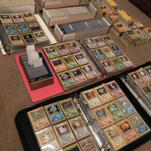 May include: A collection of Pokemon trading cards organized in a binder and several boxes. The binder is black with clear plastic sleeves and contains cards featuring various Pokemon characters. The boxes are white with clear plastic lids and contain stacks of cards.