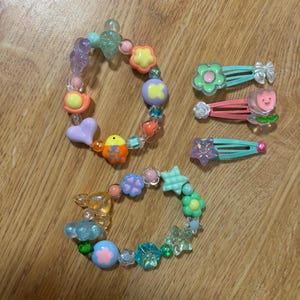 May include: Two beaded bracelets and three hair clips. The bracelets feature colorful beads in various shapes, including flowers, stars, and hearts. The hair clips are in shades of pink, green, and blue, with flower and bow designs.