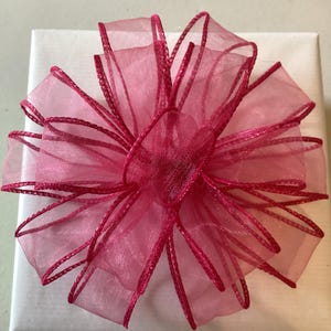 May include: A white gift box adorned with a large, bright pink bow. The bow is crafted from layers of sheer fabric, edged with a darker pink trim, giving it a textured and striking appearance. The detailed design of the bow adds a touch of sophistication to the gift.