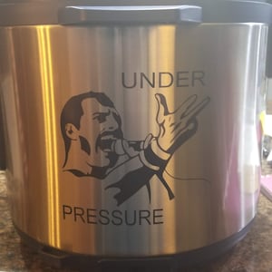 Puede incluir: Una olla a presión de acero inoxidable con un mango negro y una imagen en blanco y negro de una persona cantando con el texto "UNDER PRESSURE" en la parte delantera.