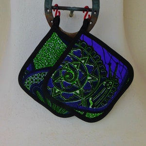 May include: Two square pot holders with black trim hang from a rusty horseshoe. One pot holder has a green and blue spiral design, the other has a green geometric pattern. The pot holders are made of fabric.