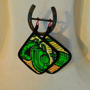 May include: Two green and yellow pot holders with a black border and a graphic of headphones hanging on a rusty horseshoe.