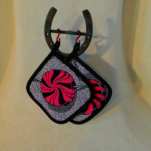 May include: Two black and white pot holders with a pink and black swirl design hanging on a metal horseshoe hook.