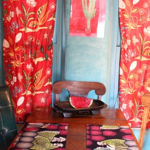 Puede incluir: Una mesa de comedor de madera con dos manteles individuales rectangulares con un gallo verde y un patrón circular morado. Un cuenco de madera tallada contiene una rodaja de sandía. Cortinas florales rojas cuelgan tras una pared azul con un cactus.