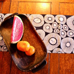 Puede incluir: Un cuenco de madera contiene una rodaja de sandía y dos naranjas. El cuenco se encuentra sobre un camino de mesa con estampado en blanco y negro, sobre una mesa de madera. La sandía es roja con semillas negras, y las naranjas son de color naranja brillante.