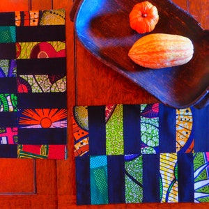 May include: Two colourful fabric placemats with a patchwork design of black, green, yellow, orange, and red. The placemats are on a wooden table with a wooden bowl and two orange gourds.