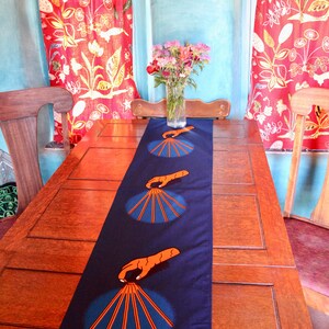Puede incluir: Un camino de mesa azul oscuro con un patrón repetido de manos naranjas sosteniendo un abanico de palos naranjas. El camino de mesa está sobre una mesa de madera con dos sillas a cada lado.