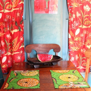 Puede incluir: Una mesa de comedor de madera preparada para dos con manteles individuales estampados en verde y amarillo, cubiertos de plata y un cuenco de madera oscura con una rodaja de sandía. La mesa está flanqueada por cortinas rojas con flores.